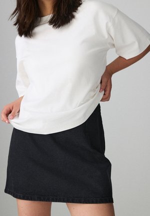 Woman wearing a plain white short-sleeve shirt and a black denim skirt, standing against a neutral gray background.