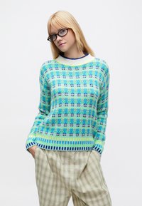 Maglione lavorato a maglia in blu e verde con motivo floreale, scollo a costine e polsini a righe. Abbinato a pantaloni chiari a quadri.