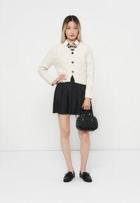 Cárdigan de punto trenzado crema, falda plisada negra, blusa blanca con cuello, bolso negro con patrón texturado y zapatos negros.