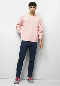 Jonge man staat tegen een witte muur gekleed in een roze kabeltrui, donkerblauwe jeans en grijze sneakers met rode veters.