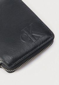 Čierna kožená peňaženka s textúrovaným povrchom, zipsovým uzáverom a vyrazeným logom CK. Okraje sú lemované; pri zipsu je viditeľné prešívanie.