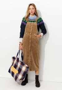 Robe salopette en velours côtelé marron avec un devant à boutons, portée sur un pull à motifs. Portant un sac fourre-tout à carreaux bleu et orange. Bottines marron.