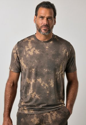 Mand med kort mørkt hår og skæg iført brun og beige t-shirt med camouflage-mønster og matchende bukser mod en ensfarvet lys baggrund.