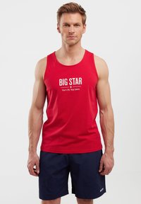 Raudona medvilninė tank top marškinėliai su „BIG STAR“ tekstu ir šūkiu, atspausdintu balta spalva; modelis dėvi tamsiai mėlynus šortus, be raštų ar papildomų detalių.