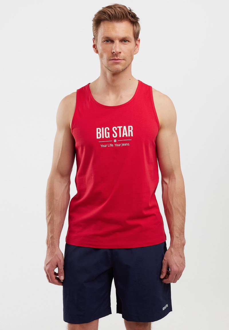 Raudona medvilninė tank top marškinėliai su „BIG STAR“ tekstu ir šūkiu, atspausdintu balta spalva; modelis dėvi tamsiai mėlynus šortus, be raštų ar papildomų detalių.