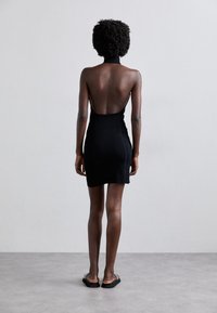 Robe noire ajustée au mini format avec un col montant et un dos nu. Tissu lisse, coutures minimales, associée à des sandales noires plates sur un fond neutre.