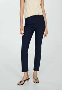 Donna che indossa pantaloni cropped slim-fit blu navy, top senza maniche beige e sandali neri con tacco basso, in piedi su uno sfondo chiaro e neutro.
