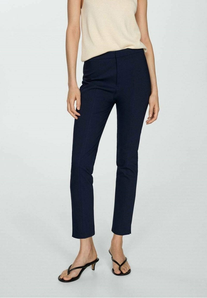 Donna che indossa pantaloni cropped slim-fit blu navy, top senza maniche beige e sandali neri con tacco basso, in piedi su uno sfondo chiaro e neutro.