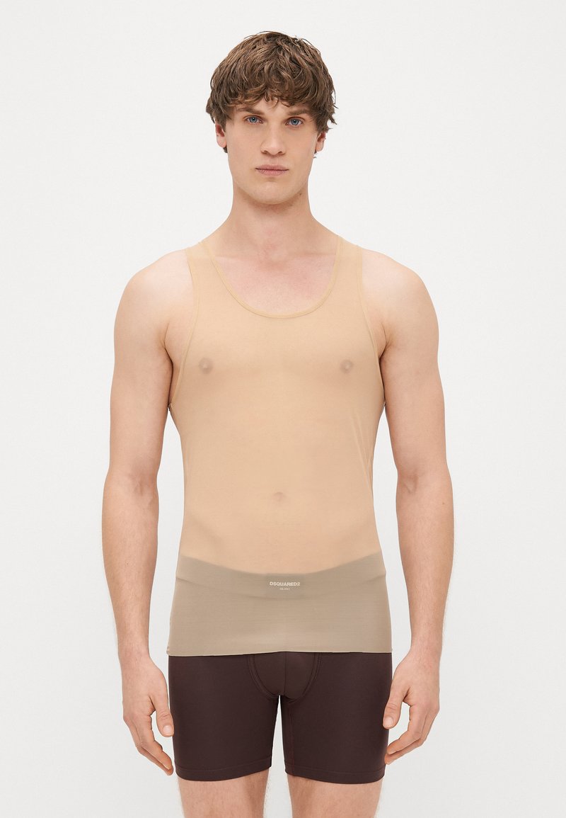 Bēša bezpaliku blūze ar plānām siksnām un gludu, elastīgu tekstūru. Valkāta virs brūnām šortiem ar zīmola elastīgu jostu.