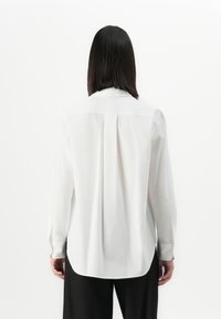 Person with straight black hair wearing a loose white long-sleeve shirt and black pants, shown from the back against a plain white background.