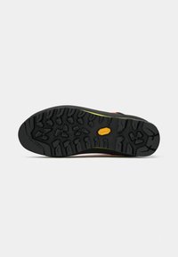 Czarna, szorstka podeszwa buta z głębokim bieżnikiem i żółtym logo "Vibram" na środku, zaprezentowana na jednolitym białym tle.