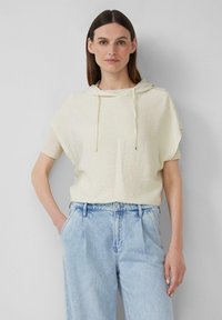 Frau mit glattem braunem Haar, die einen cremefarbenen Kurzarm-Hoodie über einem beigen Oberteil und lockeren hellblauen Jeans trägt, steht mit einer Hand in der Tasche.