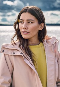 Jonge vrouw met lang bruin haar en een gele bloem achter haar oor, gekleed in een roze jas en een gele trui, kijkt zijwaarts bij het water.