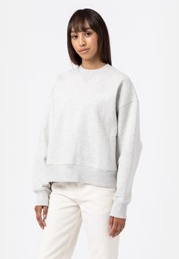 Felpa in pile grigia con spalle scese, polsini e orlo a costine, e un sottile dettaglio del logo sul lato sinistro del petto. Indossata con pantaloni chiari.