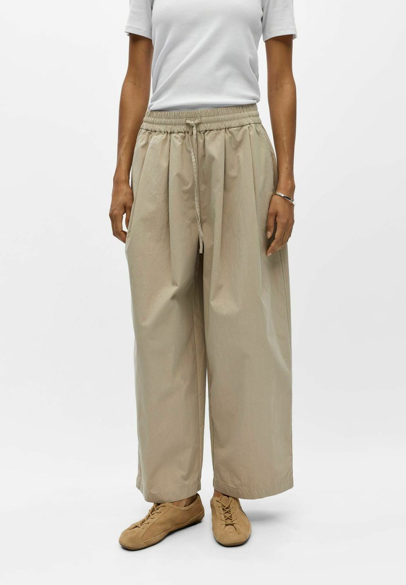 Femme portant un pantalon large beige avec cordon, des chaussures lacées couleur tan, une chemise blanche à manches courtes et un bracelet argenté au poignet droit.