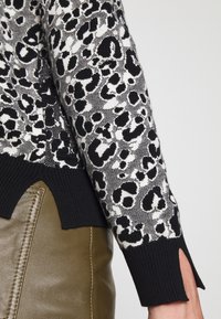 Pull en tricot avec un motif léopard noir et blanc. Il présente des poignets noirs côtelés et un ourlet courbé ; porté avec un pantalon vert olive.