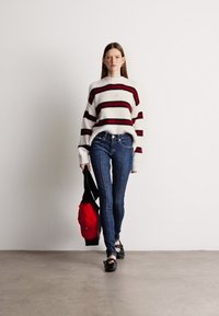 Pulôver riscado em branco, vermelho escuro e preto, combinado com jeans skinny azuis. A modelo carrega uma mochila vermelha, com sapatos pretos, num fundo neutro.