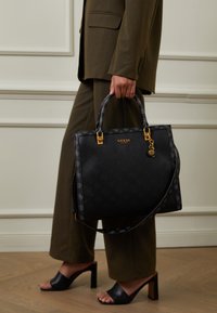 Černá kabelka Guess s texturovaným povrchem, zlatými kovovými prvky, dvojitými rukojetmi a odnímatelným popruhem na rameno. Hladká kožená materiál, čtvercový tvar.