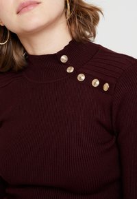 Bordowy prążkowany sweter z wysokim kołnierzem. Ozdobiony dekoracyjnymi złotymi guzikami na jednym ramieniu oraz prążkowanym detalem wzdłuż kołnierza.