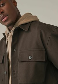 Brun vävd jacka med två framfickor, silverfärgade knappar som detaljer, som bärs över en beige hoodie. Texturerad tyg visas i närbild.
