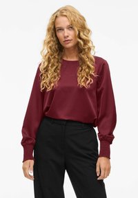 Top lungo in satin borgogna con maniche lunghe, scollatura rotonda e polsini arricciati, abbinato a pantaloni neri a vita alta. Texture liscia.