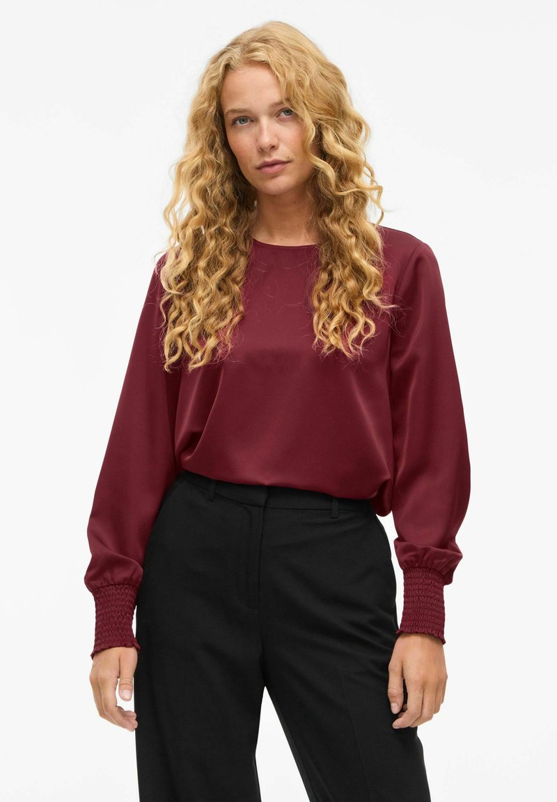 Top lungo in satin borgogna con maniche lunghe, scollatura rotonda e polsini arricciati, abbinato a pantaloni neri a vita alta. Texture liscia.