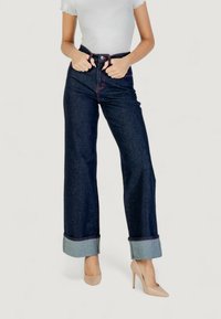 Jeans de mezclilla de pierna ancha, de un azul oscuro con costuras rojas, con dobladillos en los extremos, de cintura alta, combinados con una blusa blanca ajustada y tacones beige.