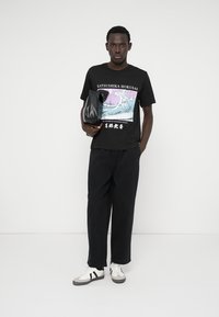 Tricou negru grafic cu un design cu valuri, asortat cu pantaloni negri lejeri. O geantă neagră este ținută într-o mână; teniși albi și gri completează ținuta.