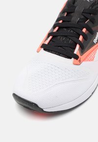 Bijela atletska cipela s crnim i koraljnim naglascima, mrežasti gornji dio, teksturirana površina, crne vezice i gumeni potplat s zaštitom za prste.