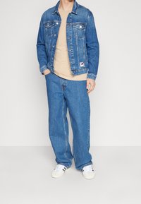 Farkkutakki, jossa on nappikiinnitys ja rintataskut, yhdistetty beigen väriseen t-paitaan ja sinisiin leveälahkeisiin farkkuihin, viimeistelty valkoisilla lenkkareilla.