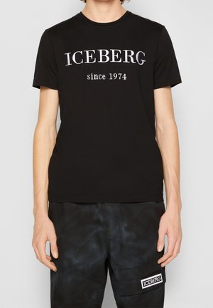 T-shirt noir avec "ICEBERG since 1974" en texte blanc, porté avec un pantalon noir comportant une poche à patch "ICEBERG".