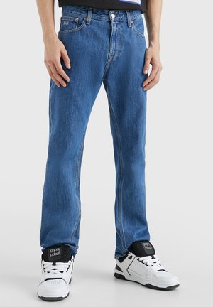 Homme portant un jean droit bleu et des baskets blanches avec des accents noirs, debout, les mains détendues le long du corps.