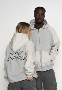 Šedá zipová mikina s krémovou kapucí a rukávy. Na zádech má vyšívaný nápis "SIMPLY APPAREL" v tučném stylizovaném písmu.