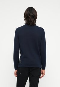 Pull en tricot bleu marine à manches longues avec un col rond, présentant une petite étiquette à l'ourlet arrière. Le matériau semble doux et texturé.
