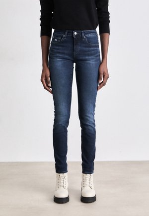 Jeans ajustados de color azul oscuro con una cintura alta, bolsillos traseros y un ligero desvanecimiento, combinados con botas de encaje color blanco roto sobre una superficie neutra.