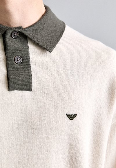 Licht beige gebreide poloshirt met een donkergroene kraag, twee knopen en een klein donkergroen geborduurd logo op de borst.