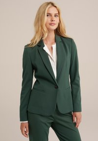 Blazer aderente verde scuro con due tasche frontali, chiusura a un bottone e rever a notched, indossato sopra una camicia bianca. Texture liscia.