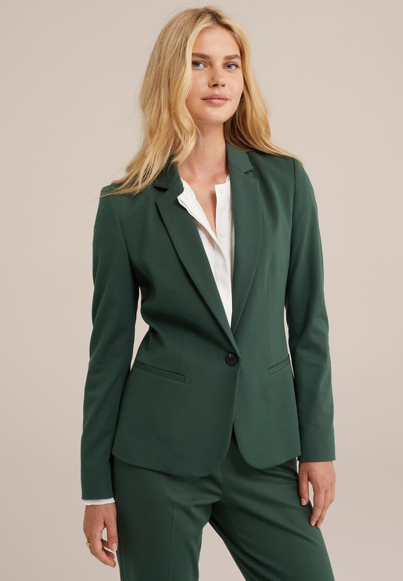 Blazer aderente verde scuro con due tasche frontali, chiusura a un bottone e rever a notched, indossato sopra una camicia bianca. Texture liscia.