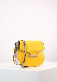 Borsa a tracolla gialla in pelle con una trama liscia, chiusura a patta, un accento in metallo di colore argento e una tracolla a fantasia bianca e nera.