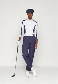 Golf ruházat cipzáras fehér és tengerészkék hosszú ujjú pólóval, tengerészkék nadrággal, fehér övvel és fehér cipővel, kiegészítve tengerészkék sapkával.