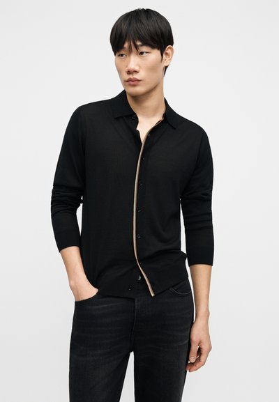 Μαύρο πουκάμισο με κουμπιά από ελαφρύ ύφασμα με μυτερό γιακά, που διαθέτει αντίθετη ρίγα περιμετρικά κατά μήκος του σκέλους με κουμπιά.