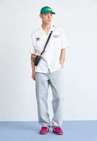 Camisa blanca de manga corta, jeans de mezclilla azul claros, gorra verde, bolso cruzado negro y zapatillas rosas con detalles en blanco. Atuendo urbano casual.