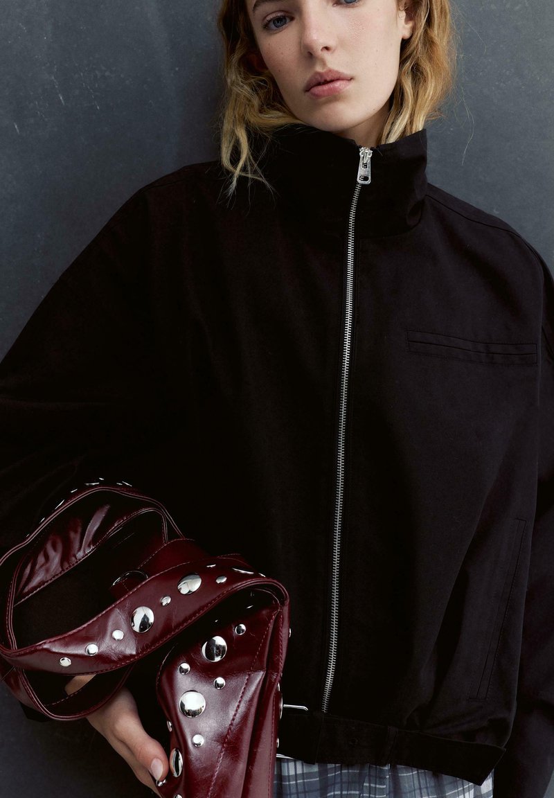 Mladá žena v černé bundě na zip drží bordó koženou tašku s velkými stříbrnými cvočky na šedém pozadí.