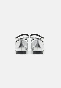 Ballerines métalliques argentées avec une bride noire à la cheville et un petit talon noir. Texture lisse avec un bout rond et des coutures soignées.