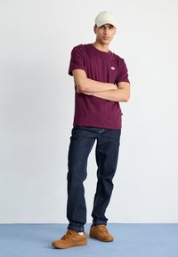 Maroon cotton T-shirt with a logo on the chest, paired with dark denim jeans. Accented by brown sneakers and a light beige cap.