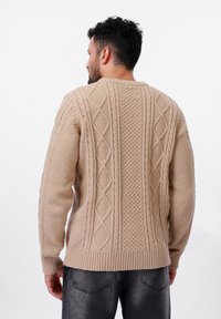 Pull en tricot beige avec des motifs texturés, doté d'un ourlet et de poignets côtelés, ainsi que de motifs en losanges et en torsades.