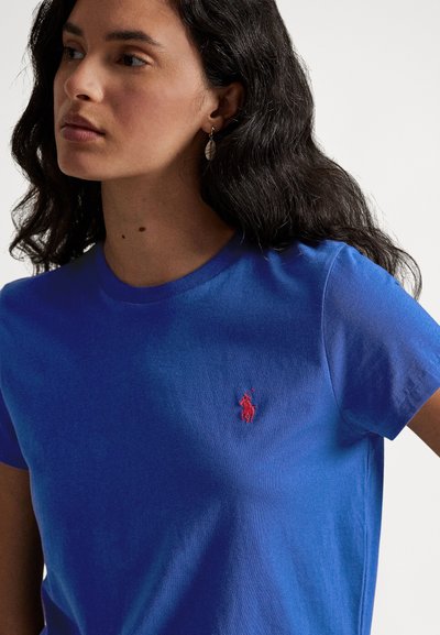 Kvinne med langt, mørkt, bølget hår som har på seg en blå T-skjorte med en rød brodert polo-spillerlogo på brystet, som ser til siden.