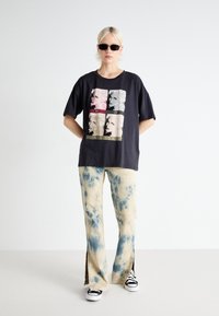 Camiseta negra de gran tamaño con un estampado gráfico de cuatro paneles. Pantalones de pierna ancha tie-dye en color beige con aberturas laterales y zapatillas negras.