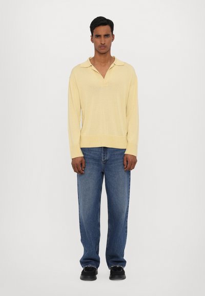 Hellgelbes Langarm-Polo mit Kragen, kombiniert mit blauen Jeans in gerader Passform. Lässige Passform mit gerippten Bündchen und Saum. Schwarze Schuhe.