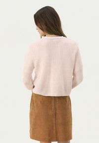Jasnoróżowy sweter dziany o luźnym kroju, prążkowanej teksturze i skróconej długości, zestawiony z brązową zamszową spódnicą. Prosty design, bez wzorów.